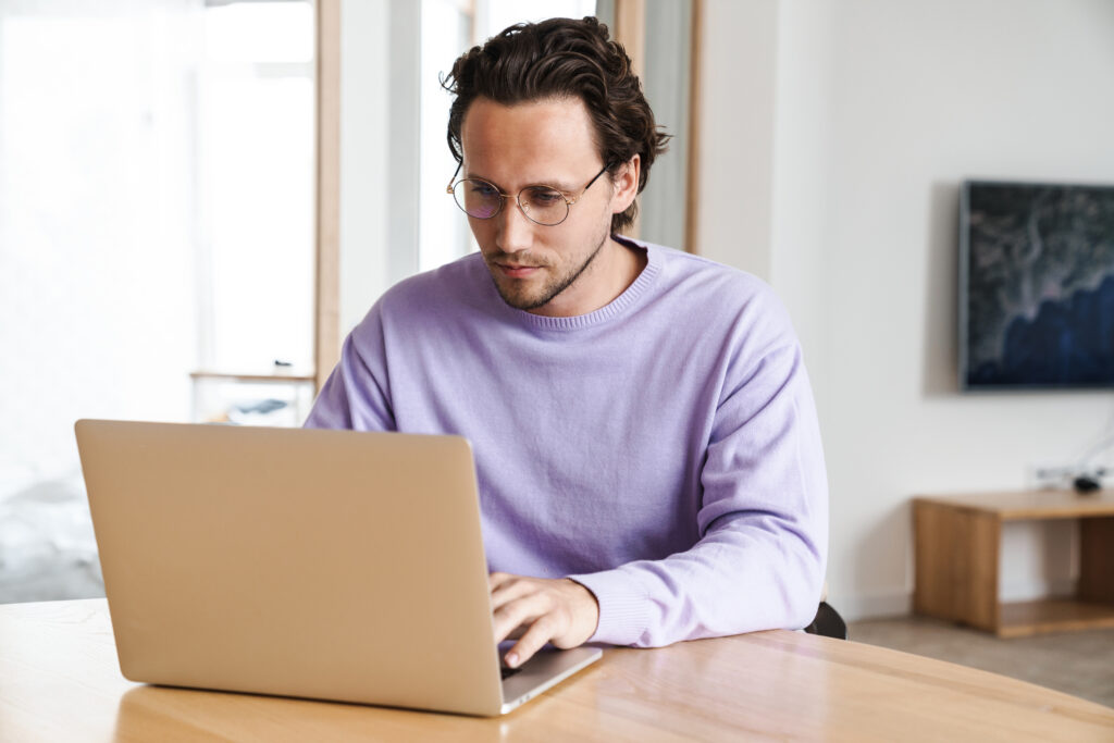 Concentrated young man sitting at the kitchen table, working on laptop computer